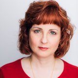 Photo of light-complexioned woman with green eyes and red hair. She is wearing a red top and a gold necklace.