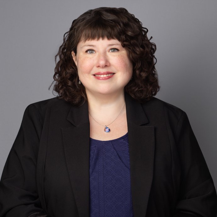 Woman with dark curly hair in black blazer and navy top smiling.