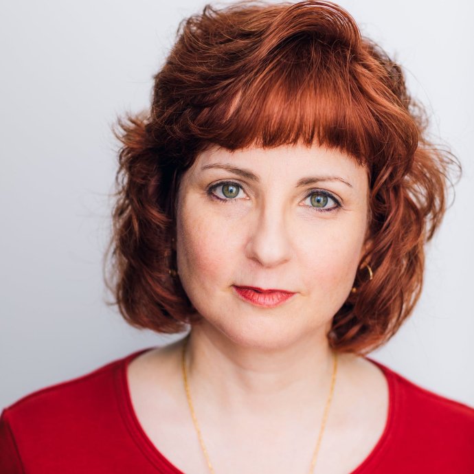 Photo of light-complexioned woman with green eyes and red hair. She is wearing a red top and a gold necklace.